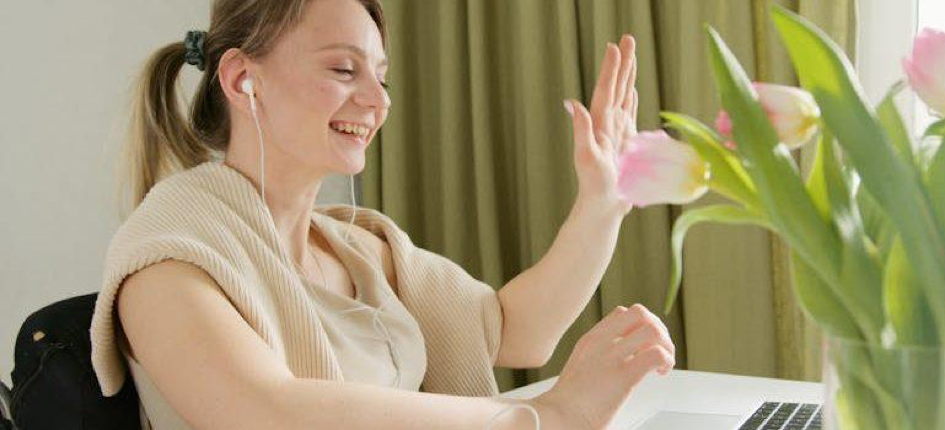 a woman with earphones smiling and waving while using her laptop